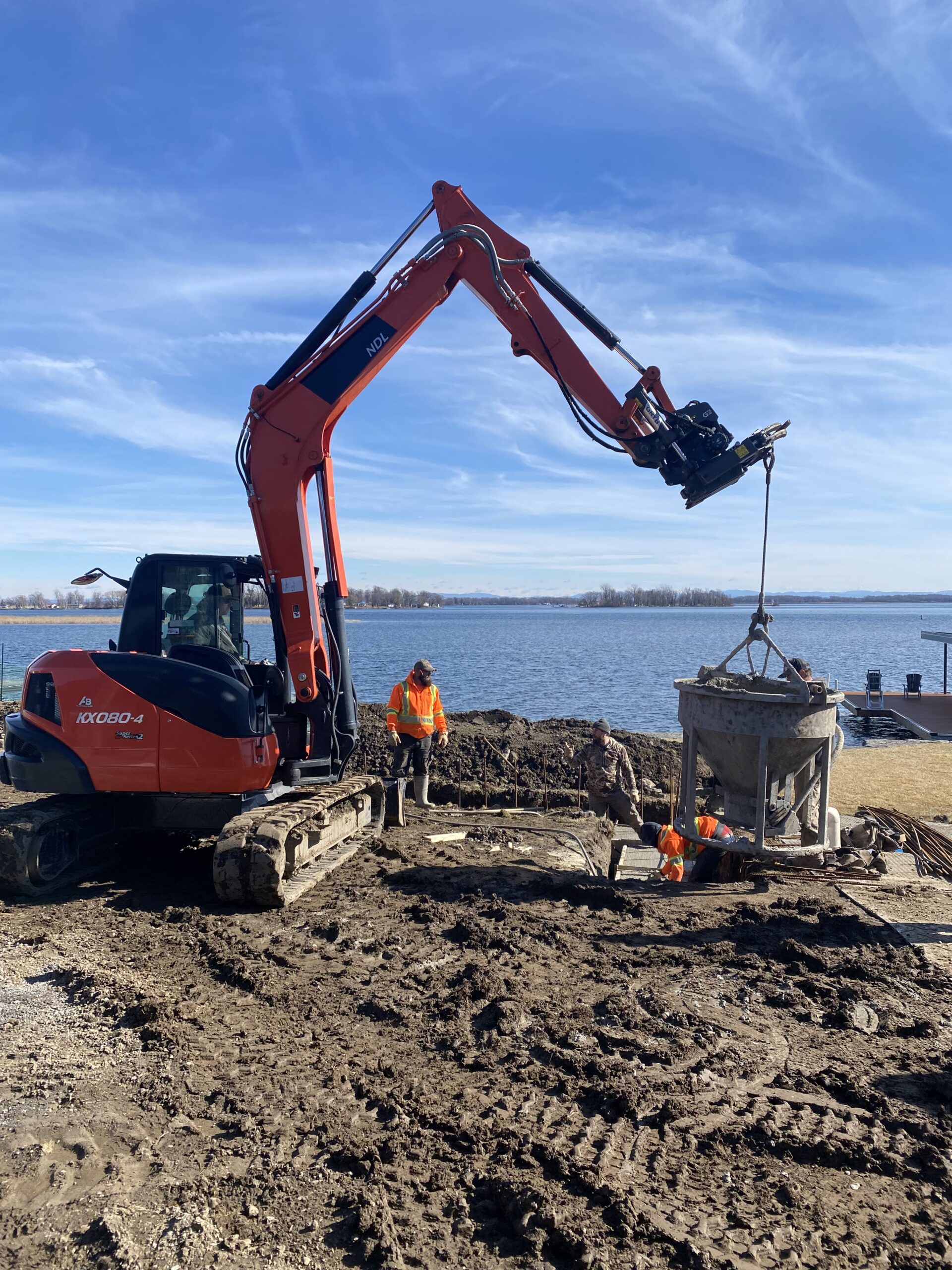 Excavator working on lakeside construction site with workers.
