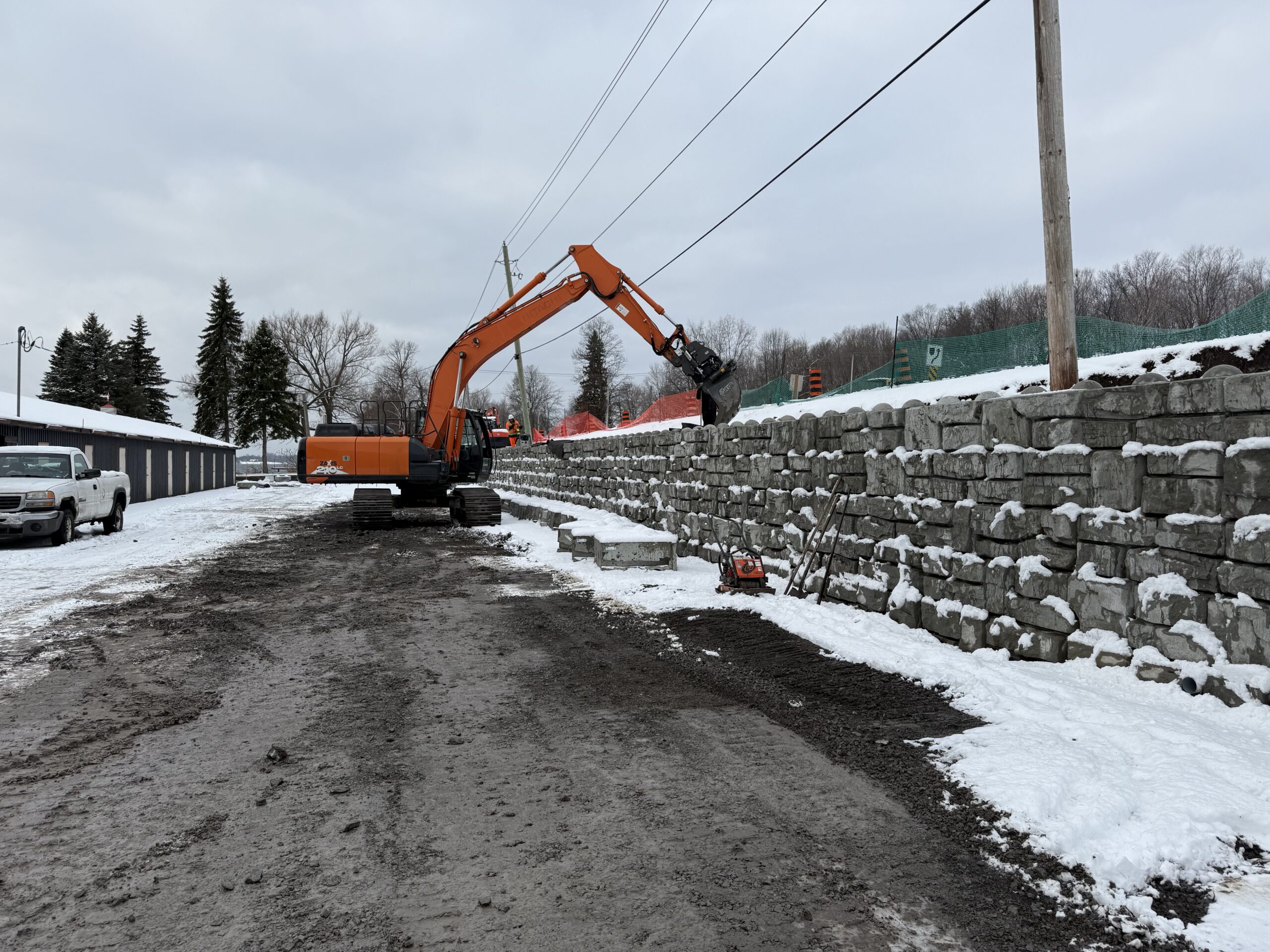 Excavator working beside snowy stone retaining wall.